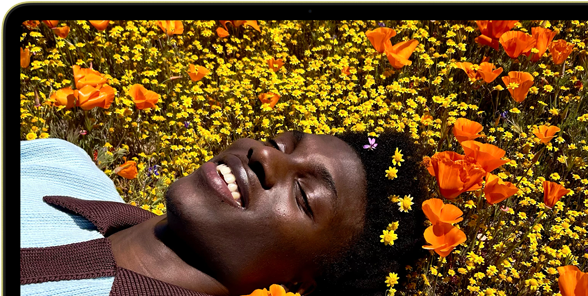 MacBook Neo, Farbe Zitrus, das Display zeigt eine Person, die in einer Blumenwiese in der Sonne liegt und lächelt