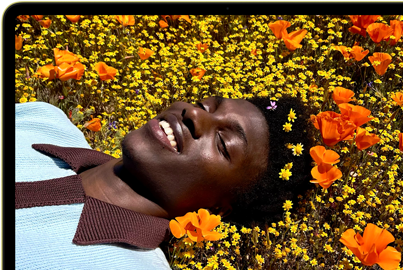 MacBook Neo, Farbe Zitrus, das Display zeigt eine Person, die in einer Blumenwiese in der Sonne liegt und lächelt